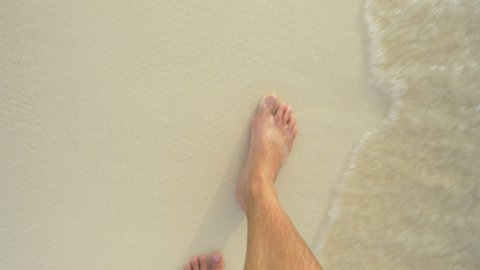 SLOW MOTION, POV: Looking down at your bare feet as you walk along the white sand beach washed by crystal clear ocean water. First person view of a man on vacation walking down a tropical sandy shore.