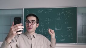 Teacher using his smartphone mobile device video chatting to explain to his students mathematical equations on a blackboard due to Online Schooling