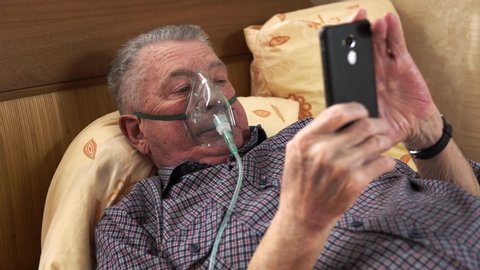 The man in breathing mask is laying at the bed and looking at the phone. Old man is using nebuliser and pushing the screen of the smartphone. Theme of medical curation of various lung diseases.