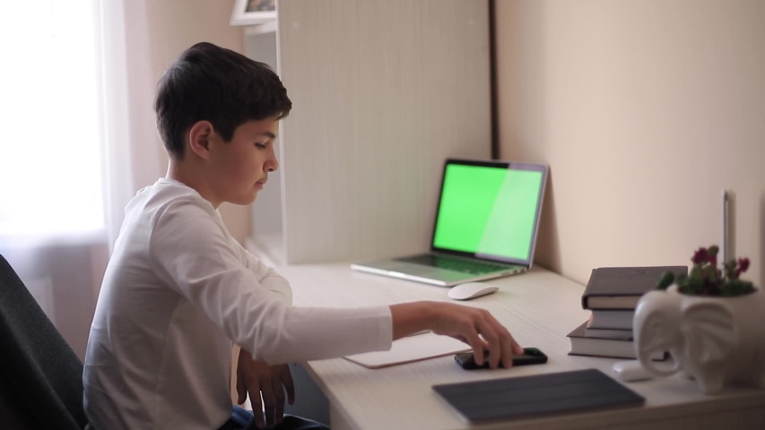 Schoolboy study at desk in his room. Boy use laptop and writing in notebook. Books and tablet on the table. Study home during qurantine. Green screen