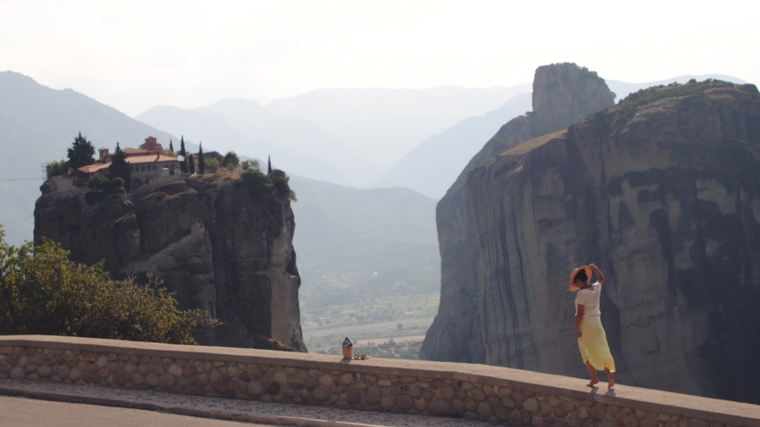 Young woman walking barefoot on a wall at Meteora rock mountains near Kalambaka, Greece. Large hat, sunglasses, fashion white dress and yellow skirt.