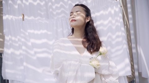 Beautiful Asian woman stands in front of drying white sheets with white roses in her hands. She enjoys wearing a fine white dress. Video de stock