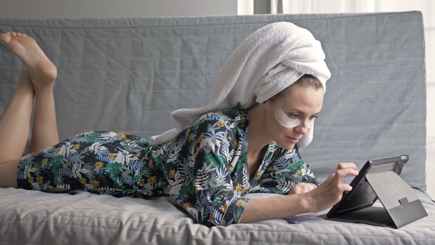 Attractive housewife after a shower is happy to shop in the online store while lying on the couch. Woman uses a laptop and a payment card.
