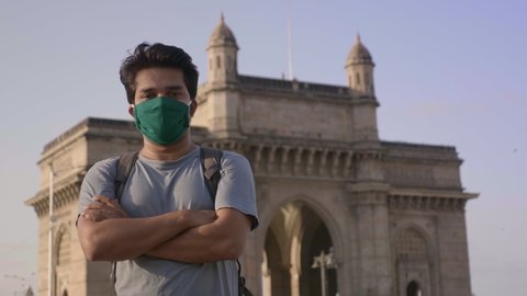 A Young and confident man wearing protective face mask looking in to the camera standing outdoors in front of famous landmark Gateway of India during lockdown amid Coroanvirus or Covid19 epidemic. 