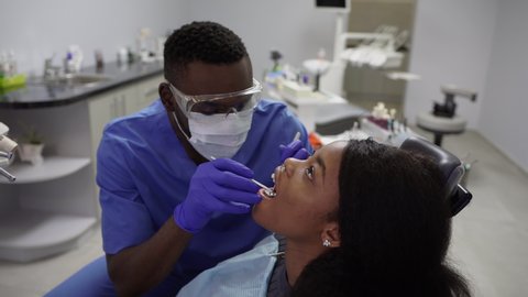 African american male dentist making treatment in modern clinic. Professional male dentist examining young african woman's teeth using special dental tools