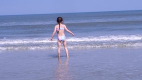 Video girl wearing in colorful hat and swimsuit enjoy playing at tropical beach. Kids enjoying warm splashing sea water on the coast. Children jump on the waves on warm sunny day on seashore.