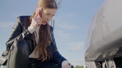 Young Cute Woman Making Phone Call After Traffic Accident. Young upset woman standing at broken car and calling for repair service or insurer. Roadside assistance concept.