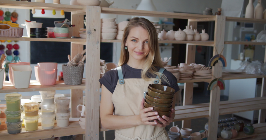 Portrait of a young smiling attractive female potter holding clay bowls. | Shutterstock HD Video #1053072584