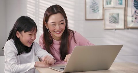 Happy mum and cute kid daughter having fun talking using learning laptop at home, smiling asian mother teach little child girl online shopping watch internet videos having fun with tech device at home