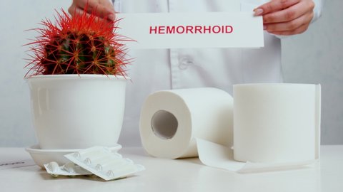Doctor putting paper with word HEMORRHOID on table with cactus, suppositories and toilet paper