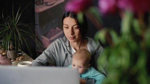 Young mother with a 6 month old baby in her arms works on a laptop while sitting at a table in the kitchen. in slow motion