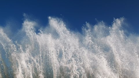 Splashes large strong waves on pier against background of bright blue sky on spring sunny day slow motion. Approaching wave with splashes of water. Strength and power of breaking wave on pier. Splash