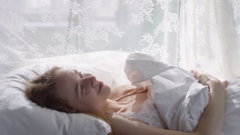  Slowmo of happy young woman in nightdress lying in bed on serene morning and smiling while enjoying sunlight shining through lace canopy