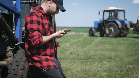 Young attractive farmer with phone standing in field, tractor working in green field in background. Smart farming using modern technologies in agriculture. Seasonal agricultural works.