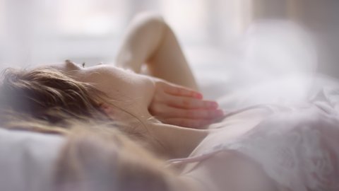  Slowmo tilt up of beautiful young woman lying in bed and enjoying tranquil morning. She is smiling and looking out window