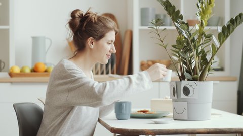 Young caucasian woman drinking tea from mug, texting on smartphone and then smiling and watering plant while sitting at table in dining room Video de stock