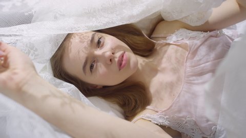  Top view of beautiful girl in nightdress lying in bed on tranquil morning and touching sheer canopy while looking at camera