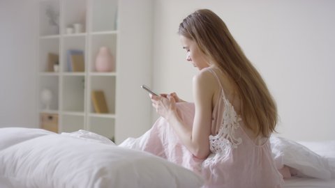  Handheld shot of young woman in pink nightdress sitting on bed on peaceful morning and typing on mobile phone