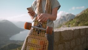 Closeup dreadlocks young woman with longboard texting on the phone, navigating map, typing message on mountain background
