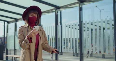 Crop view of female traveler in protective mask carrying suitcase while using smartphone near railway station. Young woman walking and looking at phone screen . Concept of coronavirus