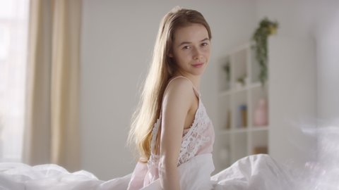  Slowmo of beautiful young woman in nightdress waking up in morning and sitting up in bed, then smiling for camera