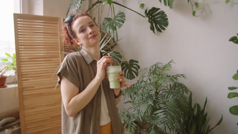Arc shot of young positive woman drinking from reusable eco-friendly cup with glass straw and then looking at camera with smile while standing in beautiful room with green houseplants