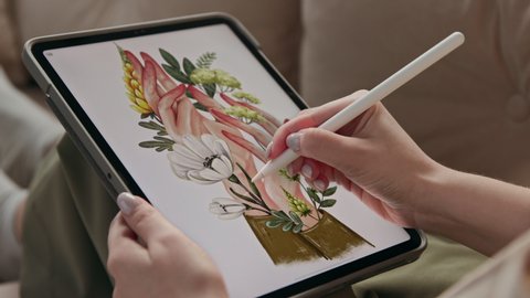 Art of young woman drawing ecological image of flowers in hands at display of digital notepad. Artist sketching colour painting by stylus at tablet computer closeup. Girl in comfort of home interior