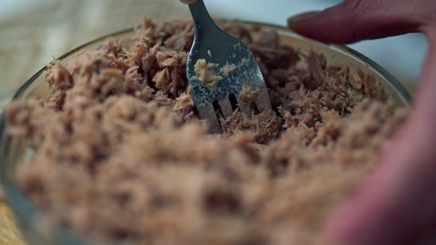 A close-up look of a woman's hand smashing/chopping tuna in a glass bowl on a wooden cutting board. Concept of crashing tuna with a fork.