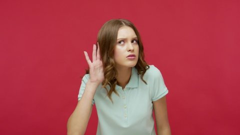 What? I can't hear you! Brunette young woman in polo t-shirt holding hand near ear to listen, suffering from hearing problems, focused on quiet conversation. studio shot isolated on red background