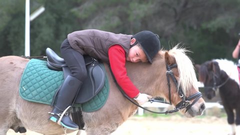 Side view of child with closed eyes in jockey costume sitting in saddle and hugging roan pony during lesson in horseback riding school