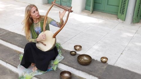 Blonde woman sitting on building stairs near singing bowls and playing lyre on street