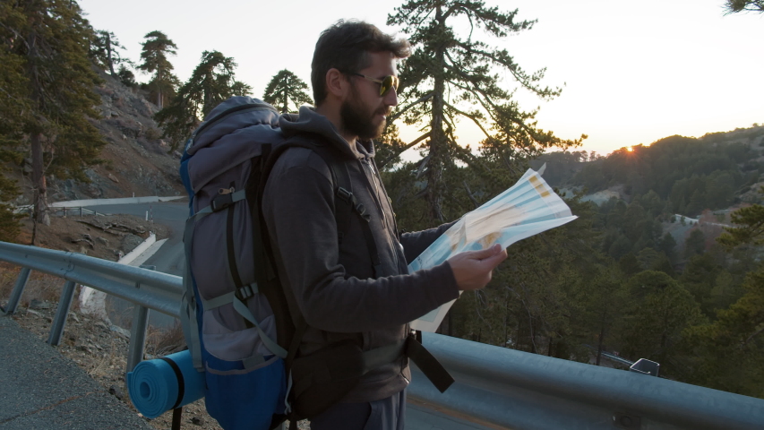 Portrait of male backpacker planning hiking trip route reading map standing at serpentine road at bottom of mountain early in morning. Traveler with map preparing to climb mountain