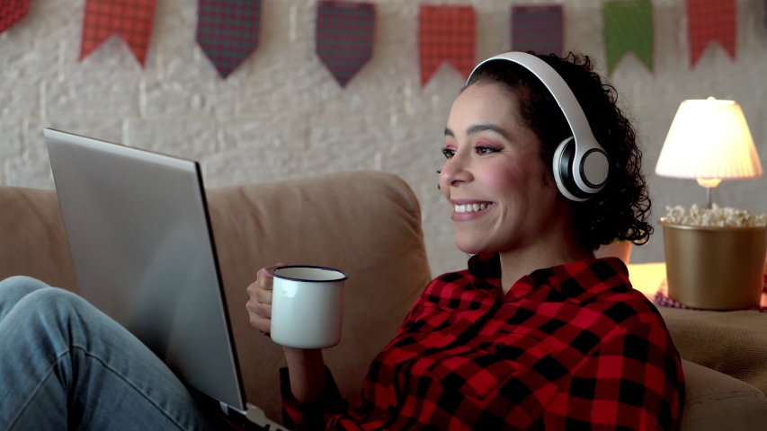 Cheerful african american woman wearing red plaid shirt drinking and watching videos online. Indoors at home living room. Traditional brazilian june festival, party, celebration concept.