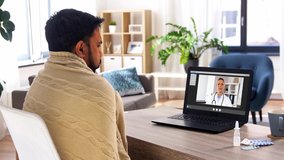 healthcare, technology and people concept - sick indian man in blanket with thermometer having video call with doctor on laptop computer at home