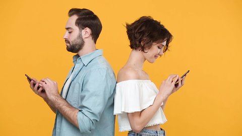 Young couple friends bearded guy spy girl in casual clothes standing back to back isolated on yellow background studio. People emotion lifestyle concept Look at gadget using mobile cell phone chatting