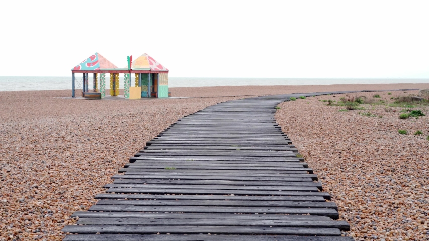 A empty wooden boardwalk along a beach looking out to sea on a cloudy day with colourful beach huts on a stone beach.