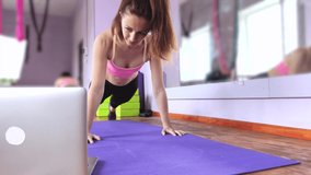Fitness and yoga trainer teaches via laptop. Wireless technology in sports. The girl shows exercises in the gym on the mat. The concept of distance learning.