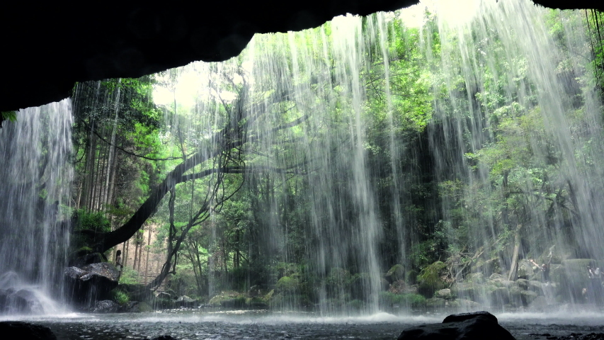 Silhouettes of people crossing in front of the camera behind an impressive waterfall in the tourist Nabegataki Falls Kumamoto Japan green nature. landscape back view shot Royalty-Free Stock Footage #1053131666