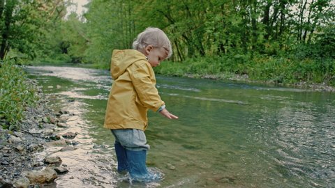 Cute baby in raincoat and rubber boots has fun by river, throws pebbles and laughs. Funny boy learns and explores natural world. Family, kids, love concept.