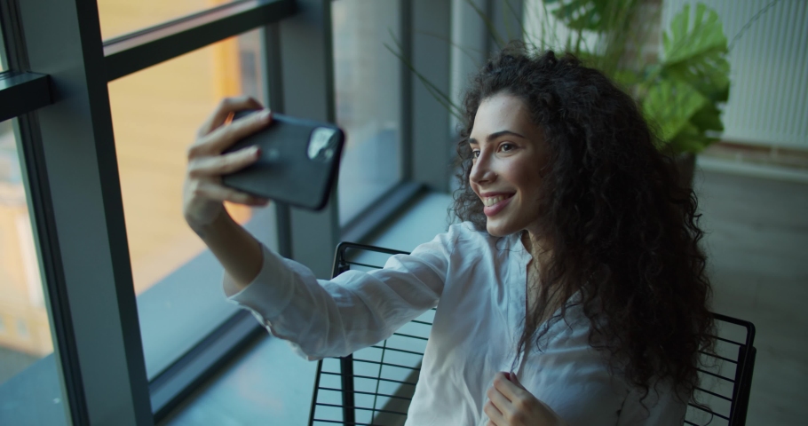 Young woman take selfie on phone while sitting on chair near window