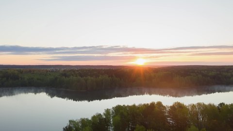 Hyperlapse drone flying over lake at sunrise. Timelapse clouds of fog above water surface. Early morning haze. Haze over river countryside. Pine trees forest reflected in water. Sun rises over horizon