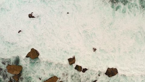 Top down aerial view from above of giant ocean waves crashing and foaming on empty sand tropical beach with big rock stones. Bird's eye aerial shot of golden beach meeting deep blue ocean water. Stockvideó