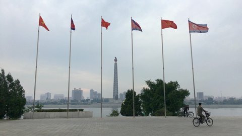 Pyongyang , Pyongyang / North Korea - 10 10 2019: National Flag of North Korea Flying Next To Worker's Party Flag. Cyclist Rides Past, Juche Tower in background. Pyongyang, DPRK