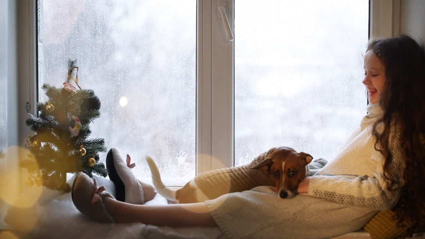 Little girl embracing her puppy dog, sitting on window and waiting Christmas.