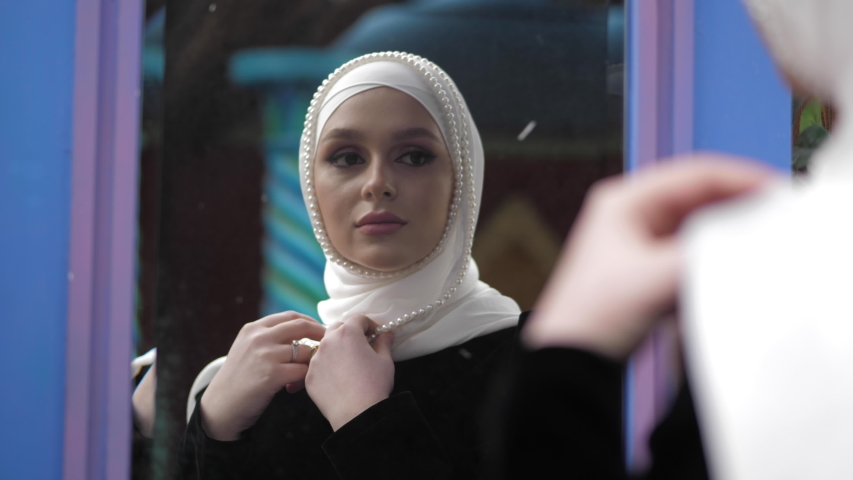 Pretty girl wearing traditional Turkish clothes and white hijab decorated with pearls looks in mirror closeup slow motion | Shutterstock HD Video #1053152168