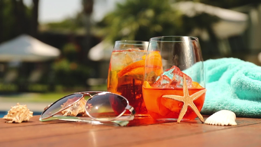 Barman is pouring champagne into aperol spritz cocktail glass with orange slices. Some seashells, towel and sunglasses near swimming pool. Vacation, summer, holiday, luxury resort concept | Shutterstock HD Video #1053160040