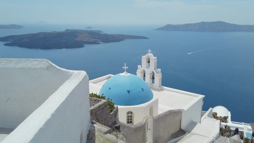 Panoramic view of Three Bells of Fira blue dommed church over caldera and vulcano island with anchored Cruiser ferry boats, Santorini, Greece Royalty-Free Stock Footage #1053160502