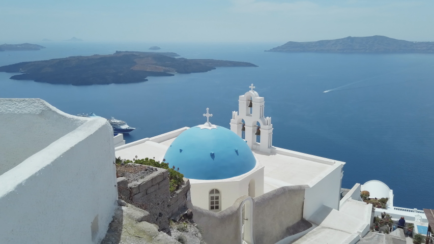 Panoramic view of Three Bells of Fira blue dommed church over caldera and vulcano island with anchored Cruiser ferry boats, Santorini, Greece Royalty-Free Stock Footage #1053160502