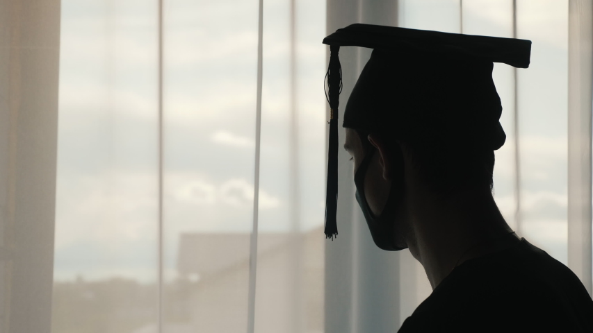 Silhouette of a graduate man in a cap and mantle and a mask on his face. Looking out the window Royalty-Free Stock Footage #1053161621