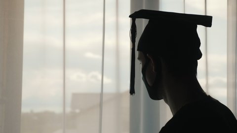 Silhouette of a graduate man in a cap and mantle and a mask on his face. Looking out the window